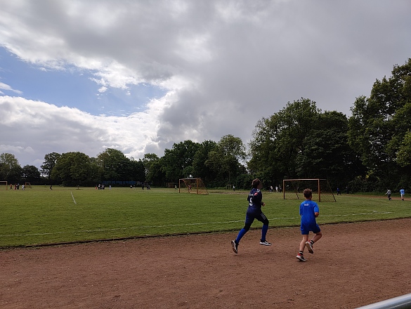 Auf dem Bild sieht man im Vordergrund zwei Kinder, die über die Rennstrecke eines Sportplatzes laufen. Im Hintergrund sind noch weitere Läufer  und Läuferinnen zu erkennen.