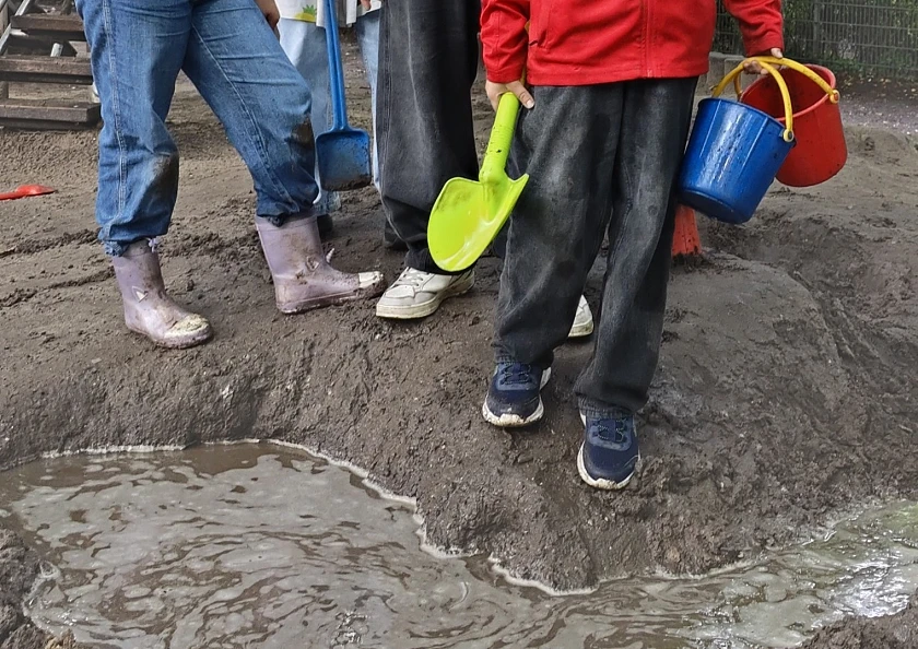 Im Vordergrund ein kleiner geschaufelter Graben mit Regenwasser gefüllt, im Hintergrund Kinder mit Schaufeln