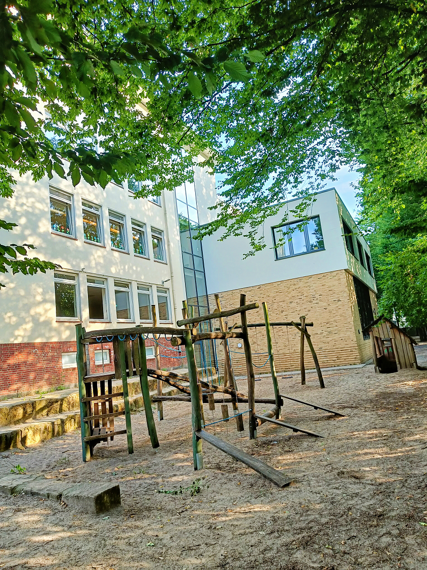 Ein kleiner Schulhof umrandet von Bäumen und dem Schulgebäude. Auf dem Schulhof sind ein großes Klettergerüst und Schaukeln zu sehen.