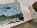 Ein fertiges Puzzle stellt den Wasserkreislauf. Ein Kind beugt sich über ein Blatt und macht sich Notizen dazu.